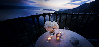 Stimmungsvoll gedeckter Tisch mit Kerzen und Blumenstrauß auf einer Terrasse mit Blick auf eine Küstenlandschaft bei Sonnenuntergang.